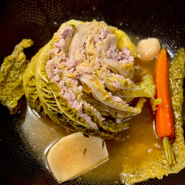Slice of stuffed cabbage with pork in a poached liquid with carrot and turnip served in a big bowl, a rustic French dish