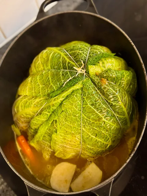 large casserole pot with a stuffed savoy cabbage tied with string and surrounded with stock and vegetables