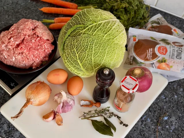 a savoy cabbage with ingredients laid out around it to make a stuffed recipe (pork, eggs, stock, onion, garlic, herbs and seasoning