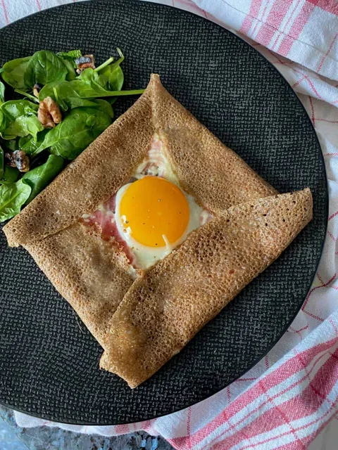 square brown buckwheat crepe pancake folded like an envelope with egg yolk in the middle surrounded with a little ham and cheese and side salad