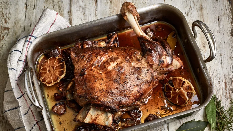 Roasted lamb shoulder with halved garlic and pan juices in a metal roasting tray.
