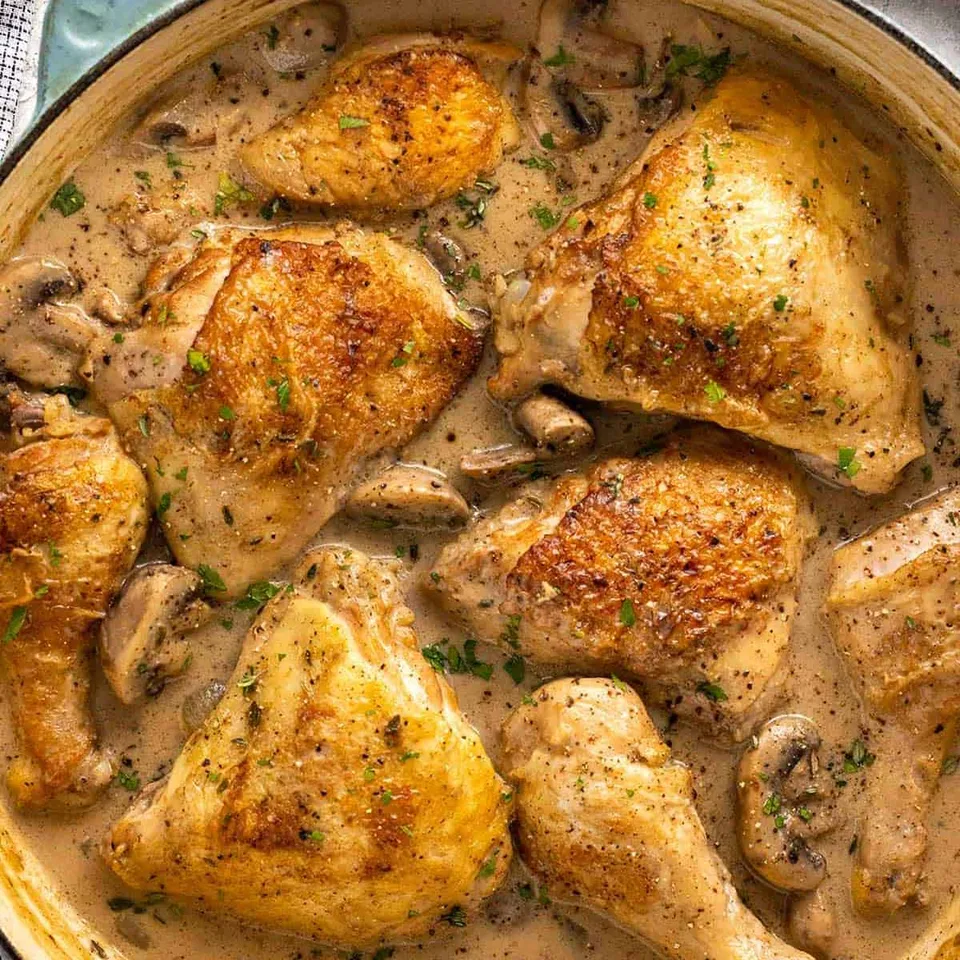 Overhead view of browned chicken thighs and sliced mushrooms in a creamy sauce.