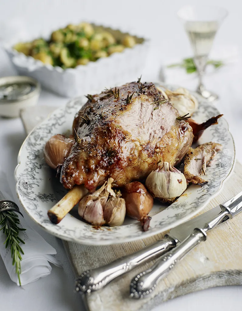 Roast leg of lamb with garlic bulbs and herbs on a serving platter.