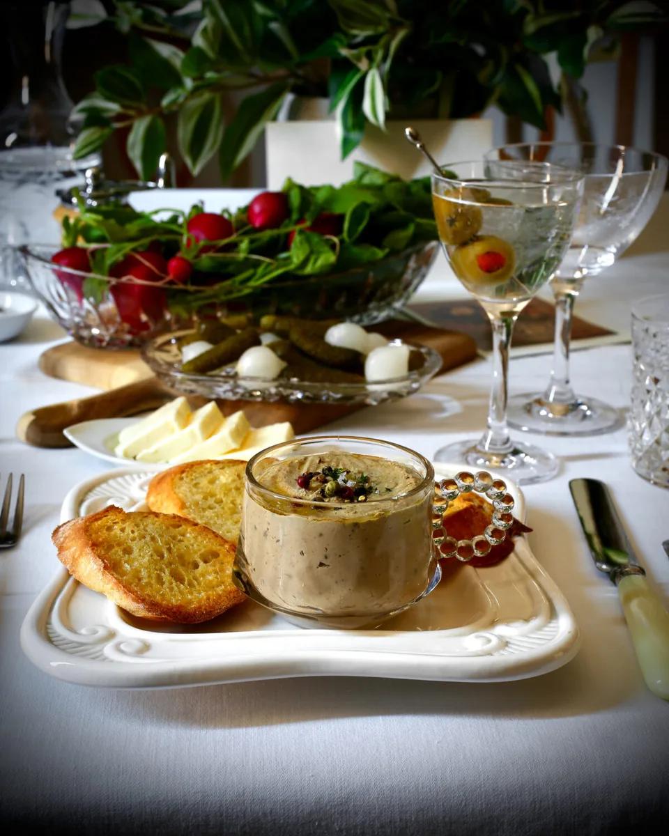 Chicken liver pate in a glass cup with toasted baguette and a martini on a table.