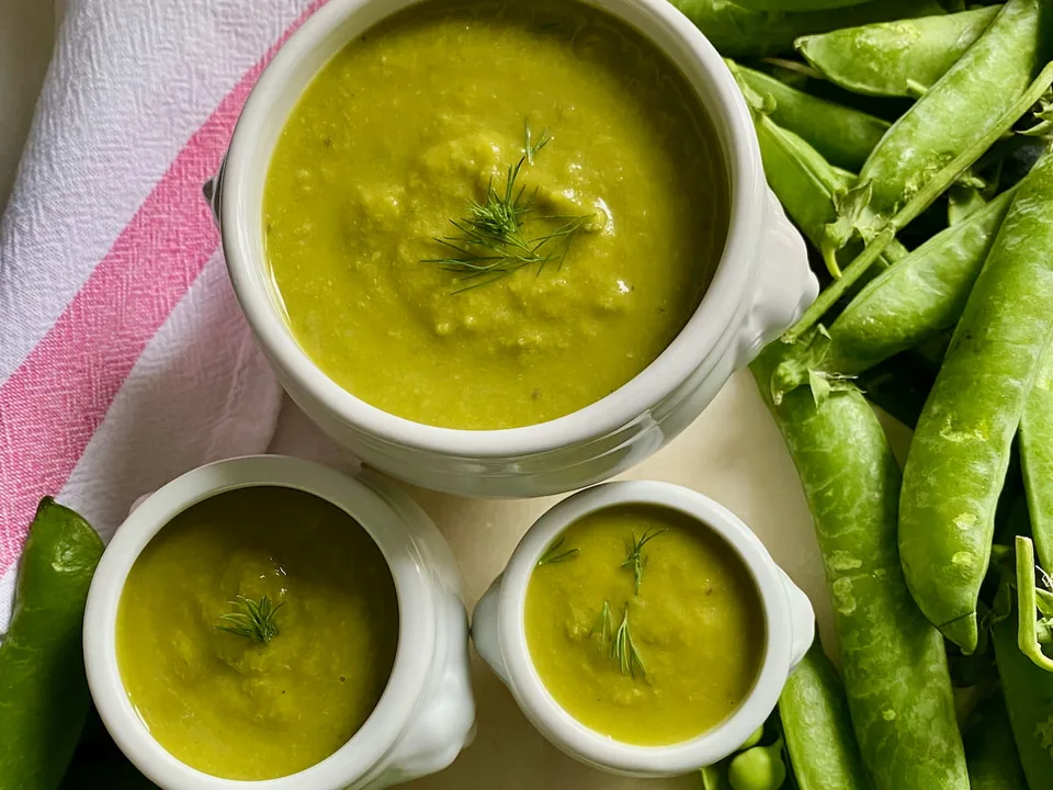 Three bowls of green pea soup with herb garnish, beside pea pods.
