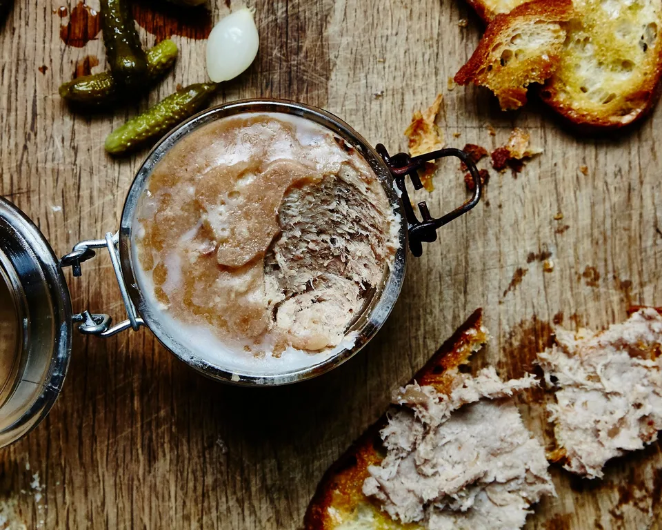 Overhead close-up of pork rillettes in an open jar with toast and cornichons on wood.