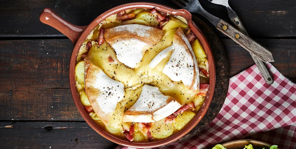 Tartiflette in a brown handled dish on dark wood, with large melted Reblochon wedges.